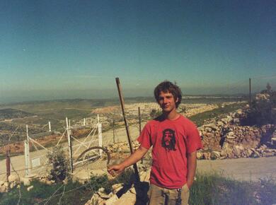 young man, graphic tshirt with a crucified Christ, holds a sickle, stands atop a hill, fences below