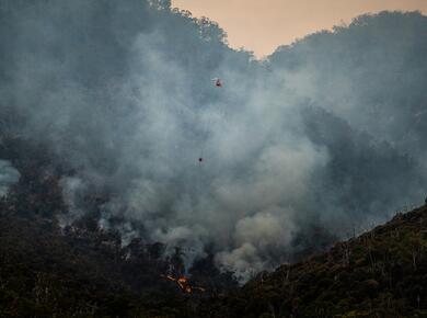 a helicopter is dwarved by a cloud of smoke from wildfires on a mountainside