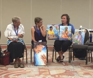 Audrey Kanagy displays three completed “Women in Conversation” decoupage paintings while explaining the project. Looking on are Nancy Heisey and Elie Soto. PHOTO CREDIT: Harriet Bicksler